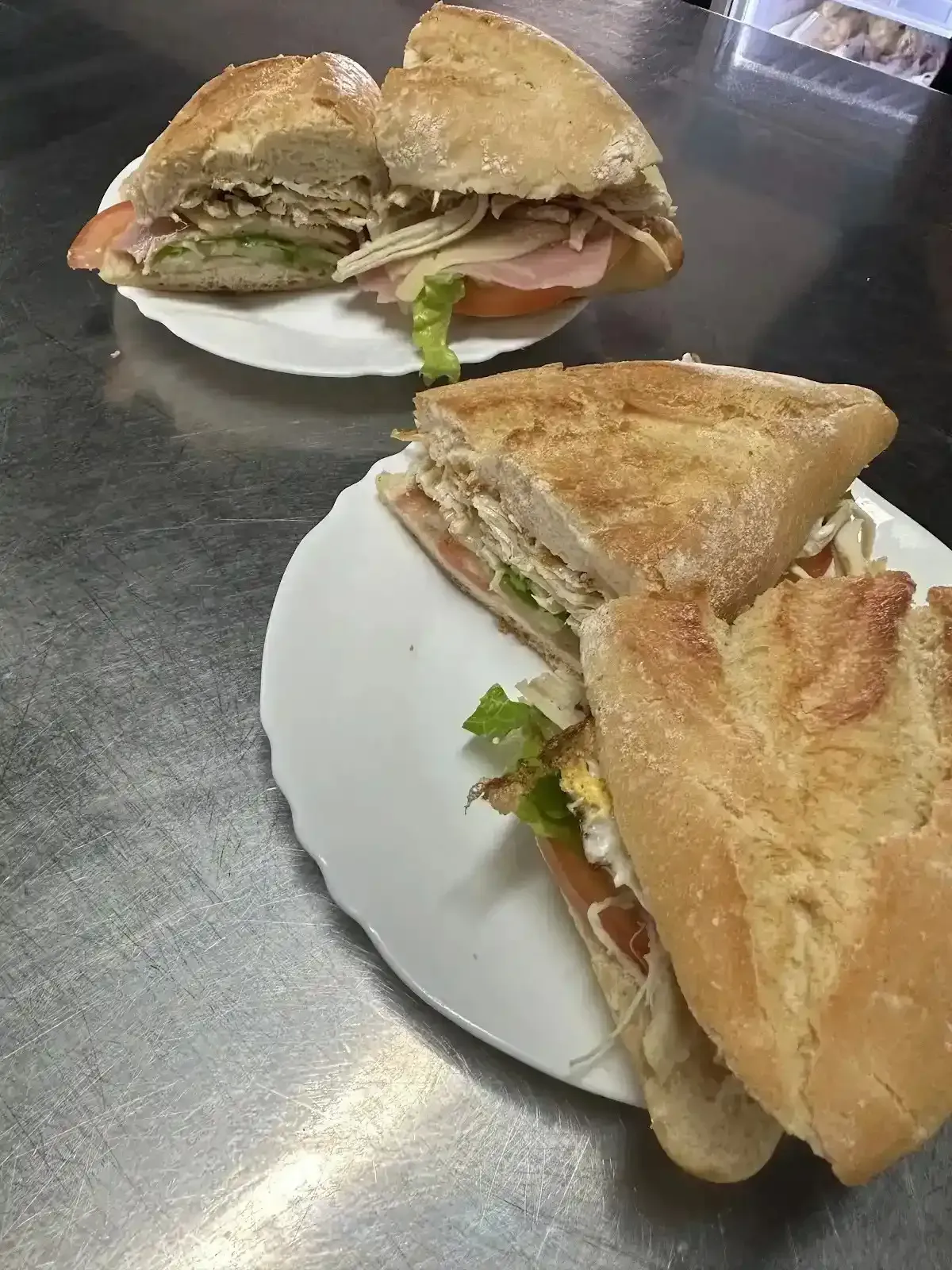 Dos mitades de sándwich en platos blancos, con lechuga y tomate sobre un mostrador de acero inoxidable.