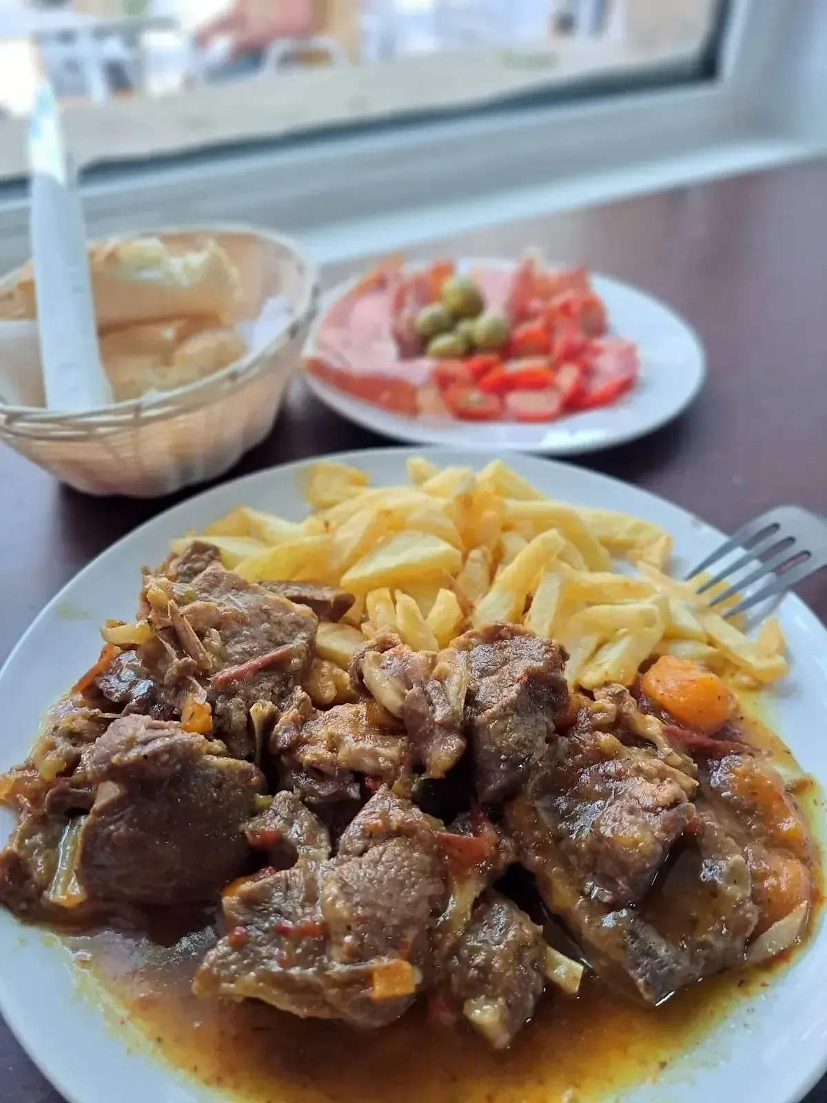 Plato de carne guisada con patatas fritas, servido con ensalada y un cuenco de salsa en una mesa.