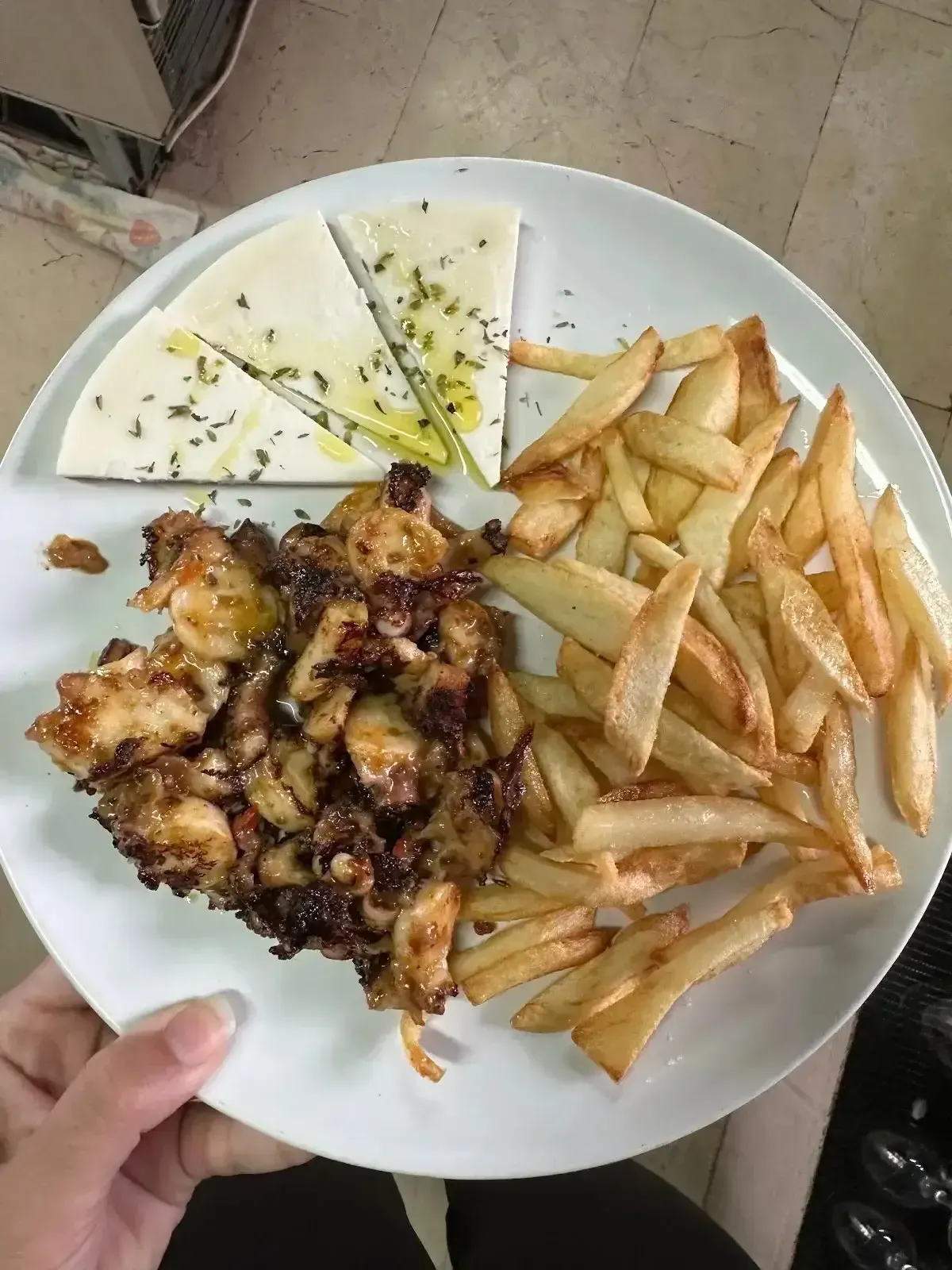 Plato con pollo a la parrilla, patatas fritas y trozos de queso blanco, sostenido en una mano sobre una mesa.