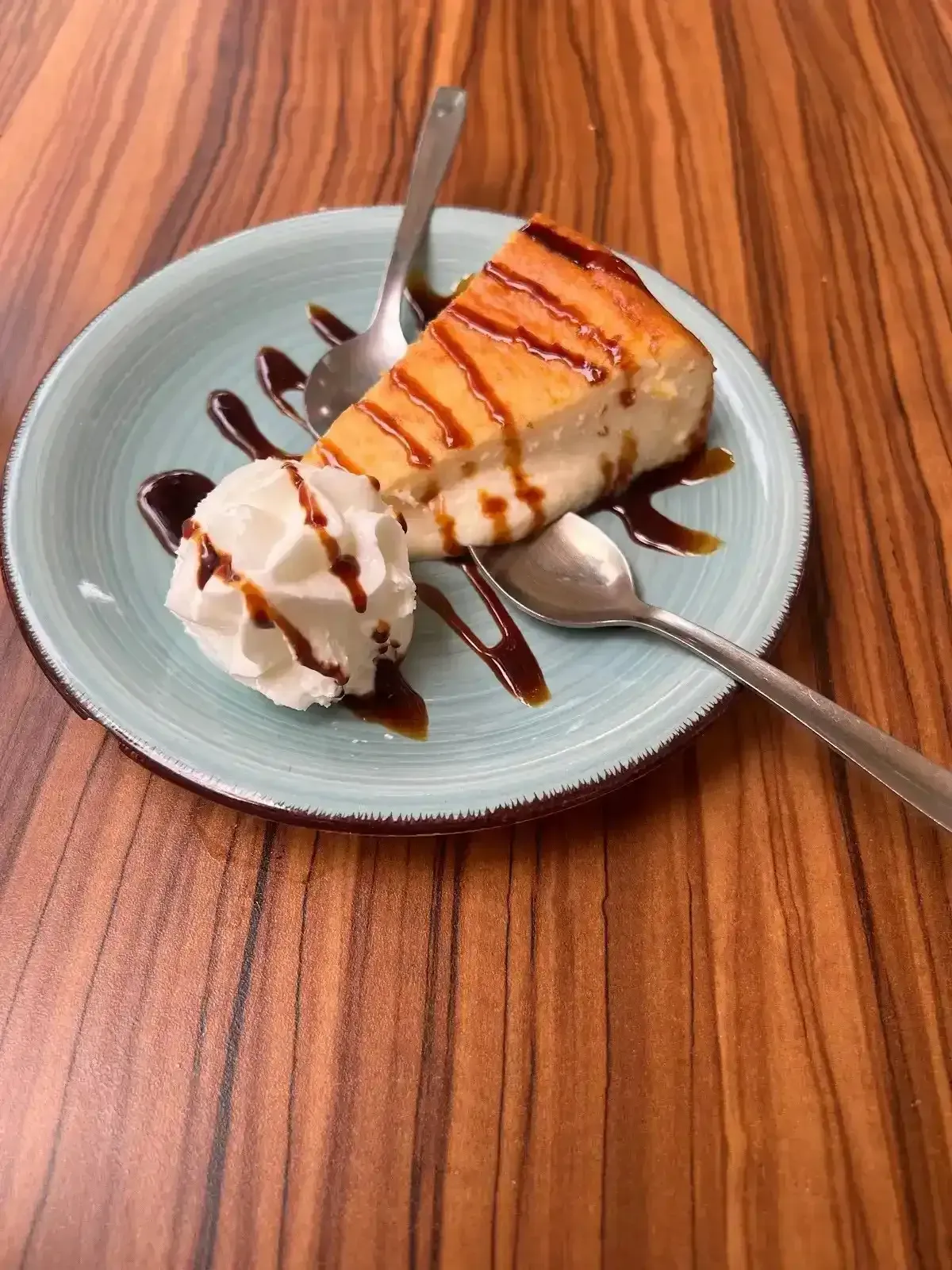 Rebanada de pastel con crema batida y sirope de chocolate en un plato azul sobre una mesa de madera.