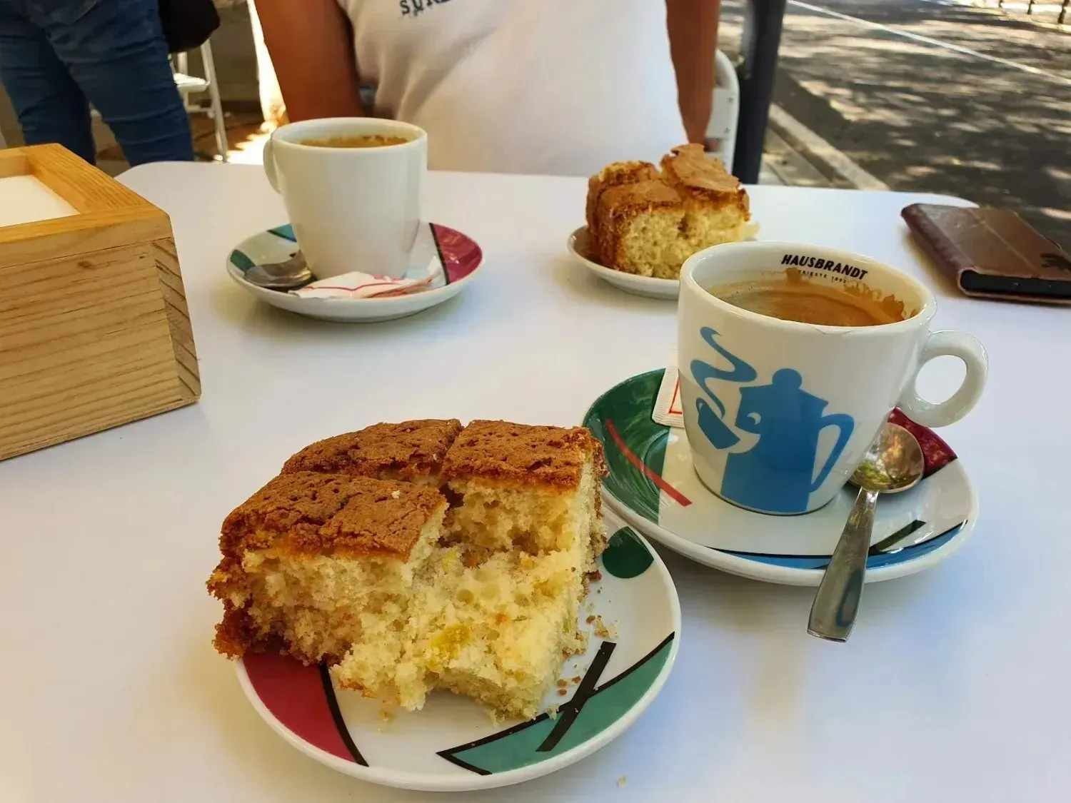 Tazas de café y porciones de pastel sobre una mesa blanca de cafetería, con gente sentada al fondo.