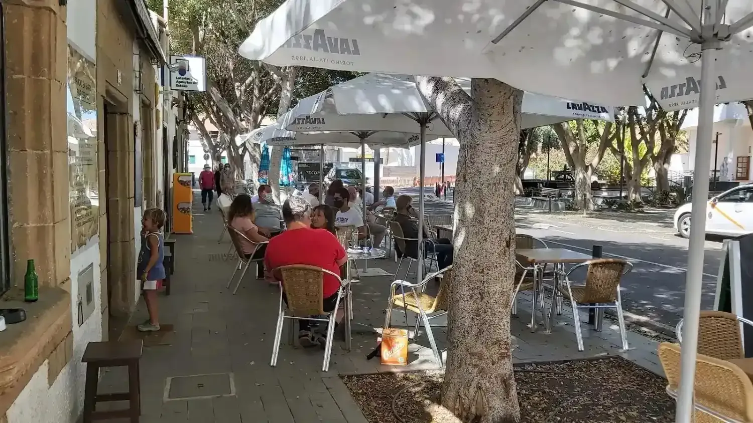 Terraza de una cafetería en una acera soleada, con sombrillas, árboles y algunas personas comiendo.