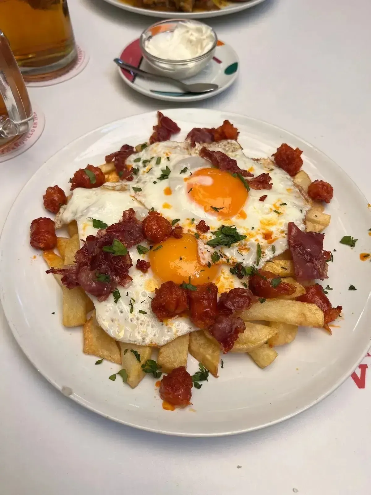 Papas fritas con extras, cubiertas con huevos fritos, trocitos de tocino y hierbas en un plato blanco.