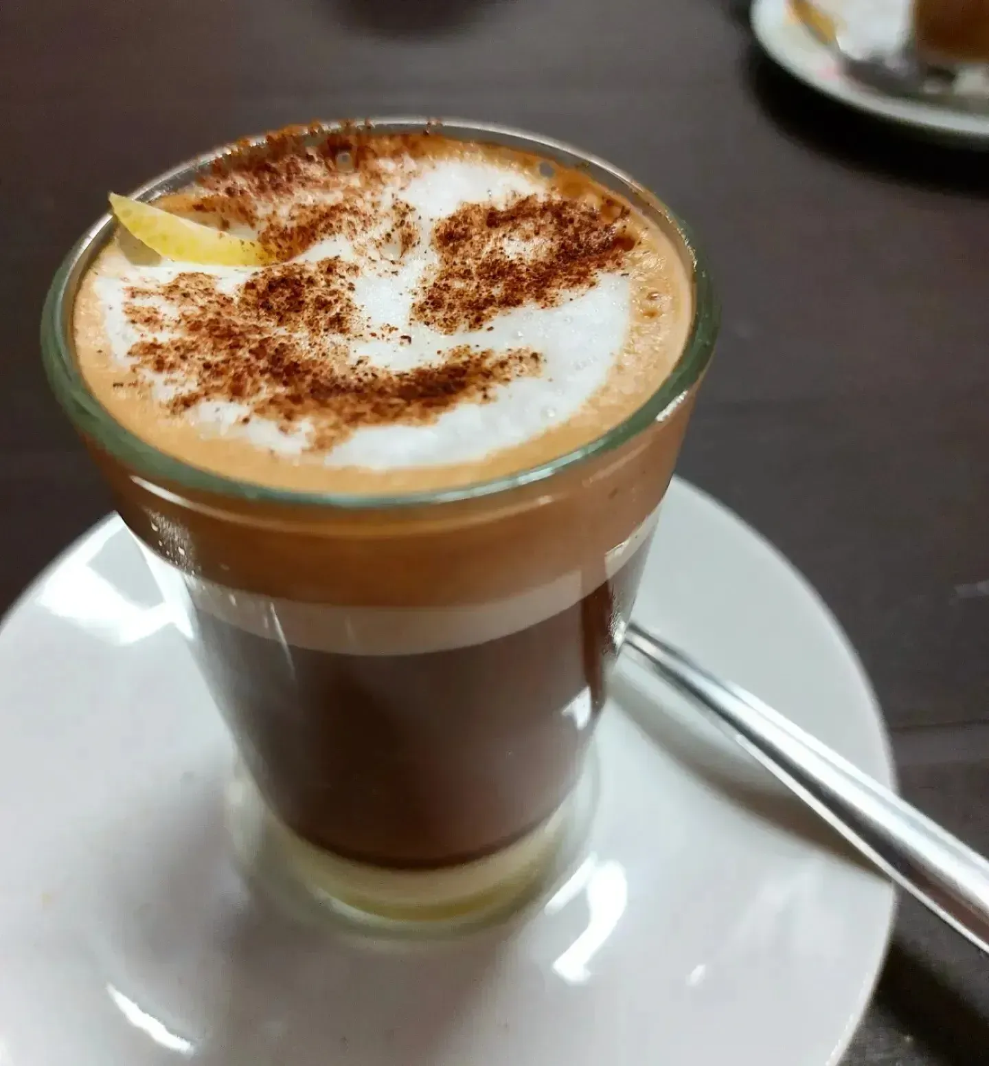 Bebida de café en capas, coronada con espuma y canela, servida en un vaso sobre un platillo blanco, con una cuchara al lado.