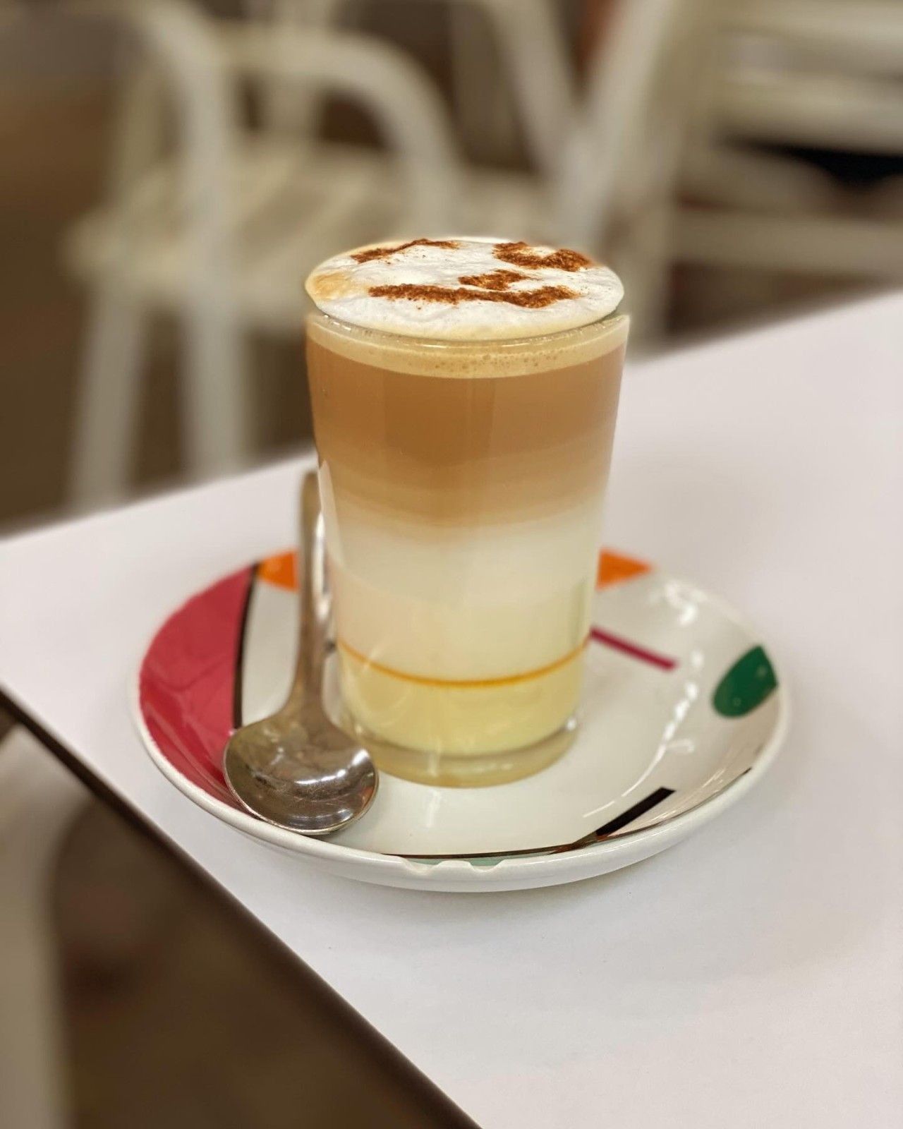 Un café con leche con espuma en un platillo, espolvoreado con canela, junto a una cuchara sobre una mesa blanca.