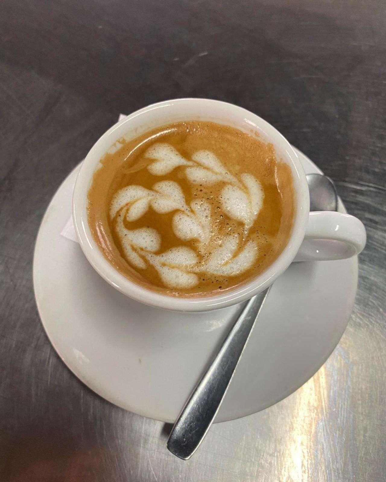 Taza blanca de café con leche decorada con dibujos de hojas sobre un platillo, vista desde arriba sobre una mesa de metal.