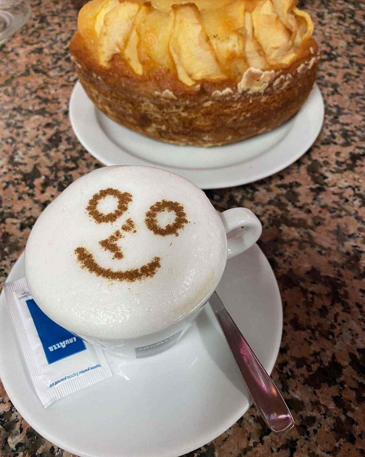 Dos bebidas de café: un capuchino con espuma y una carita sonriente, y un pastel de manzana sobre una mesa.