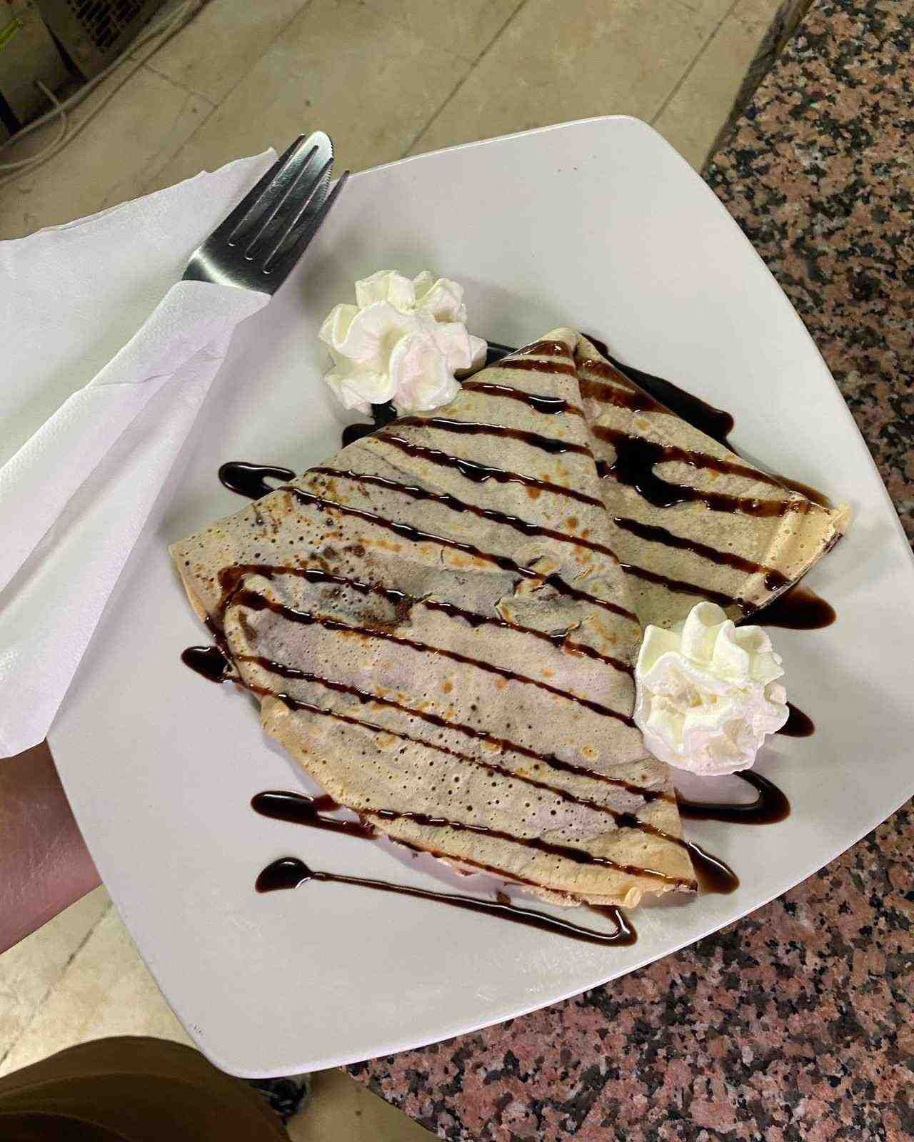 Crepe de postre bañada en chocolate sobre un plato blanco, coronada con crema batida.