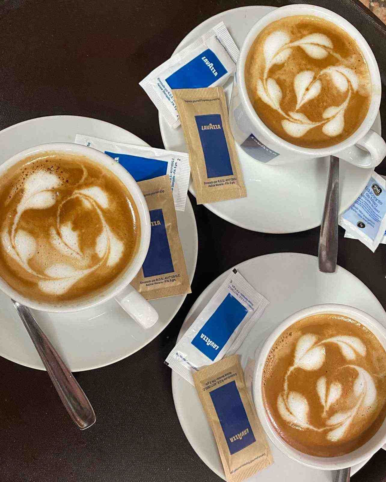 Tres tazas de capuchino con arte latte en platillos blancos y galletas sobre una mesa oscura.