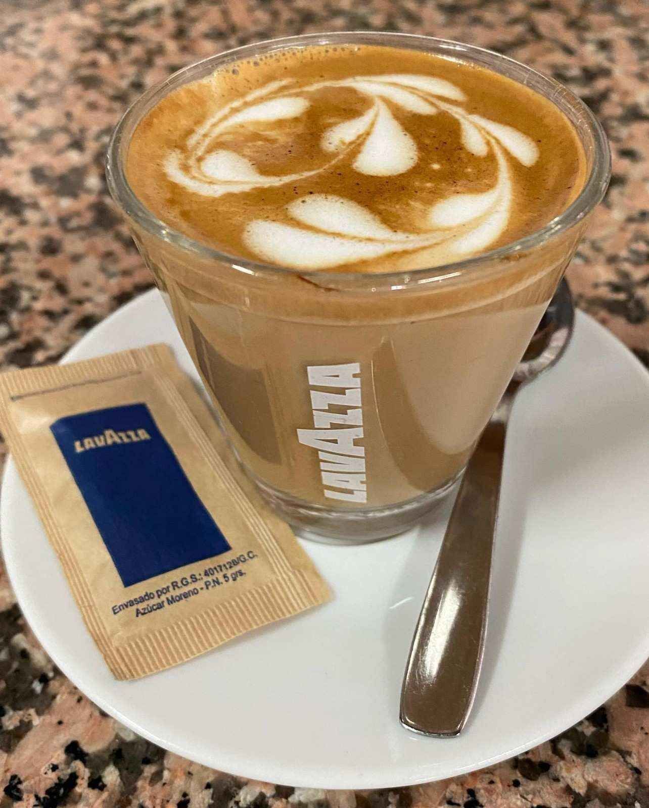 Un café con leche decorado con arte latte en un platillo, junto a una cuchara y un sobre de azúcar en una mesa de café.