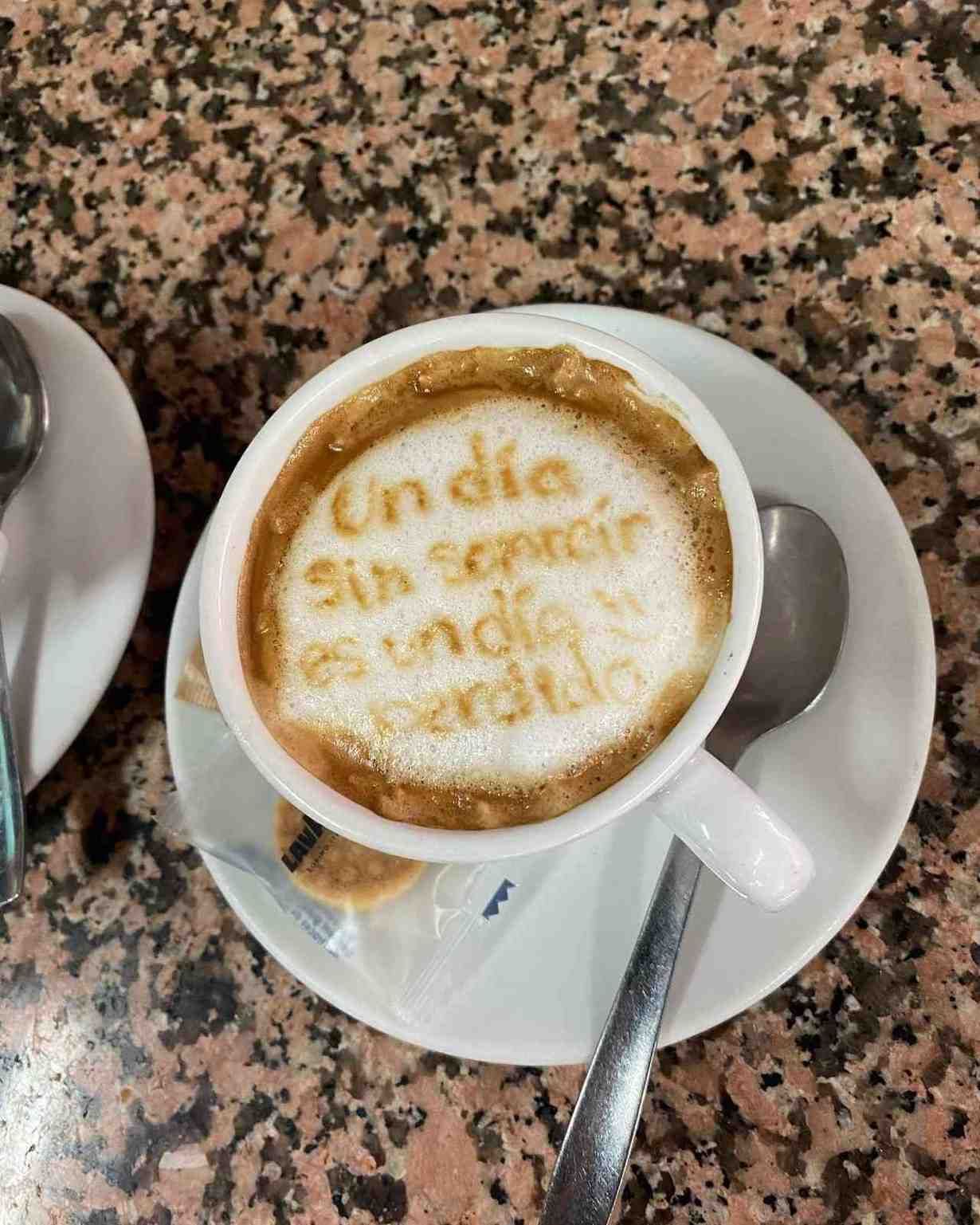 Taza de café sobre un platillo con espuma en la superficie y texto apenas visible, sobre una mesa de granito.