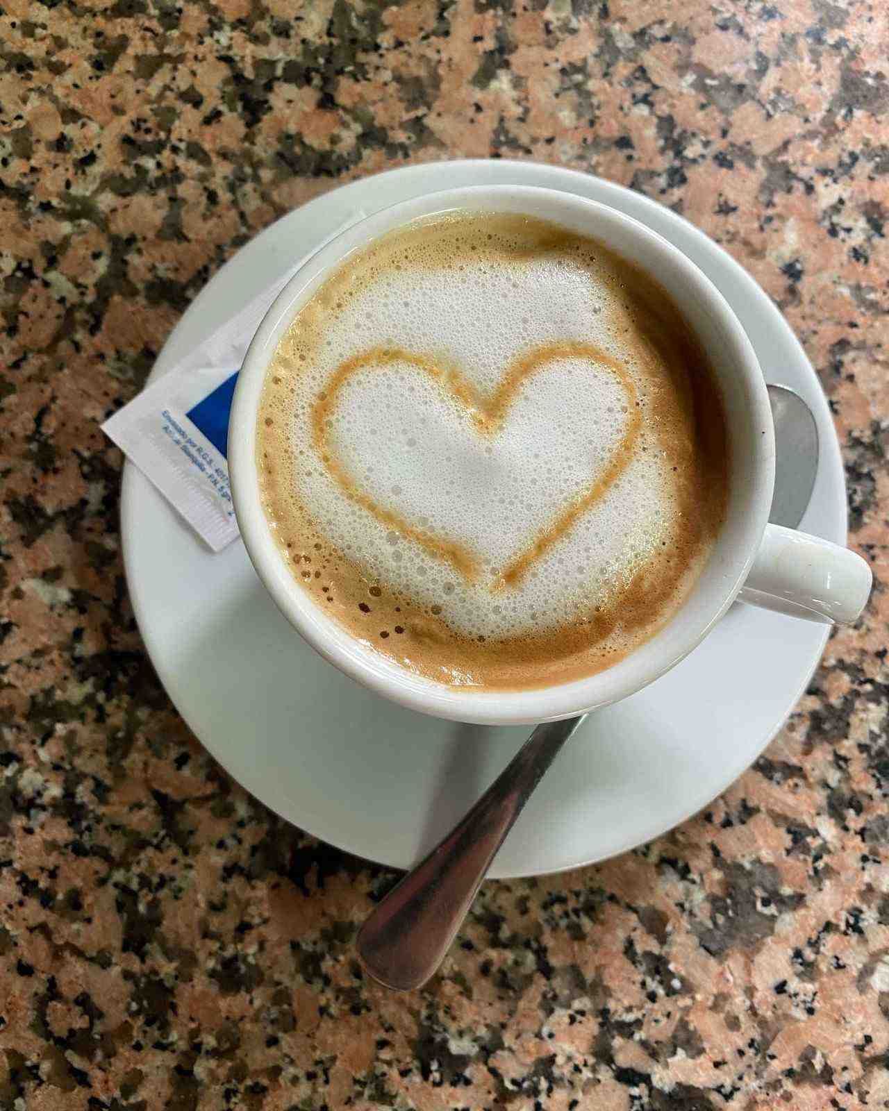 Taza blanca de capuchino con un dibujo de espuma en forma de corazón sobre un platillo, sobre una mesa de granito.