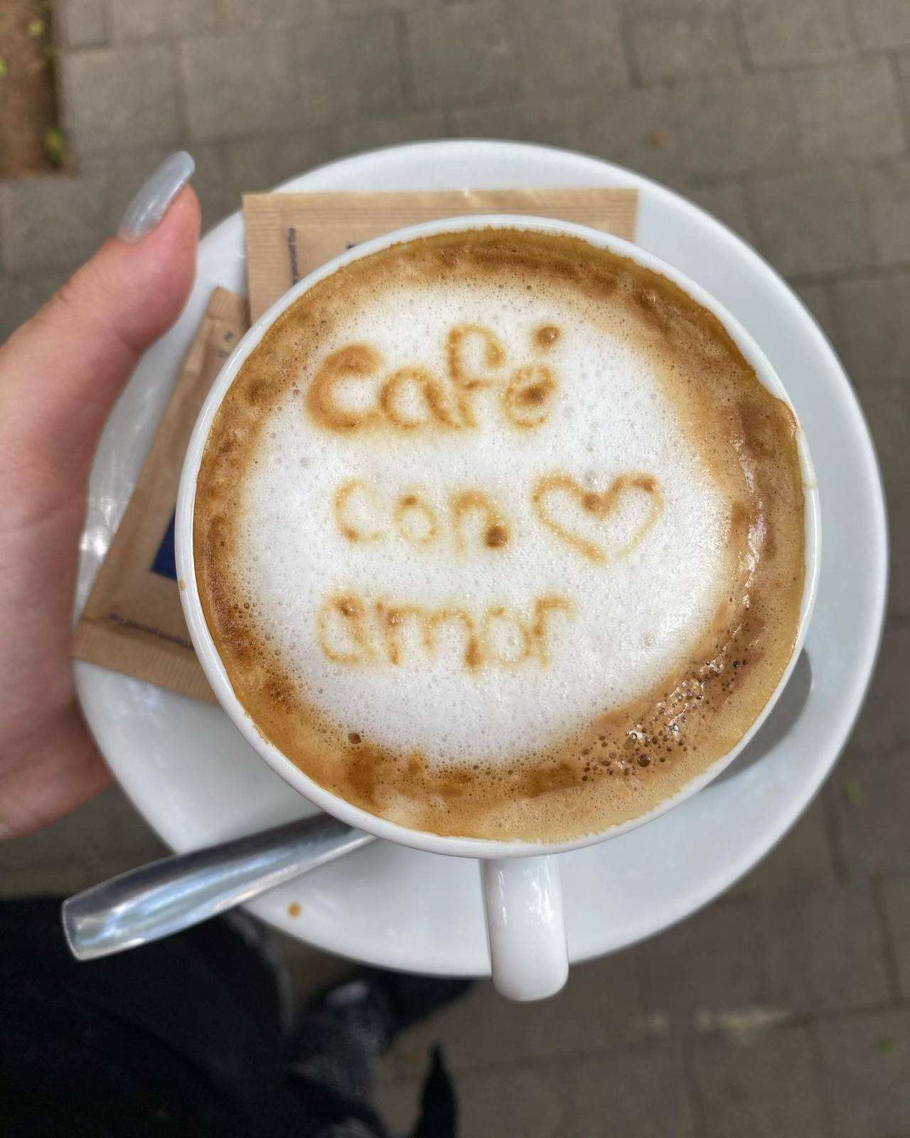 Café con leche decorado con espuma que dice “café con amor” y un corazón, sostenido sobre un platillo en una mesa.