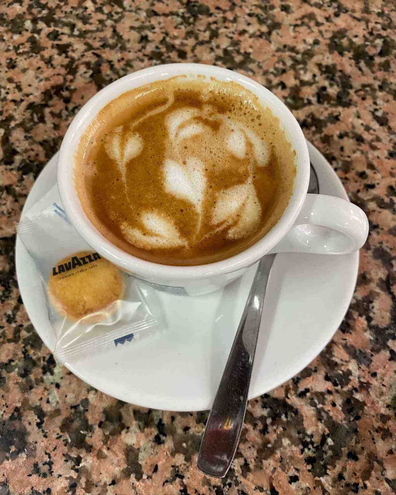 Una taza de capuchino con arte latte en un platillo, una cuchara y una galleta envuelta sobre una mesa de granito.