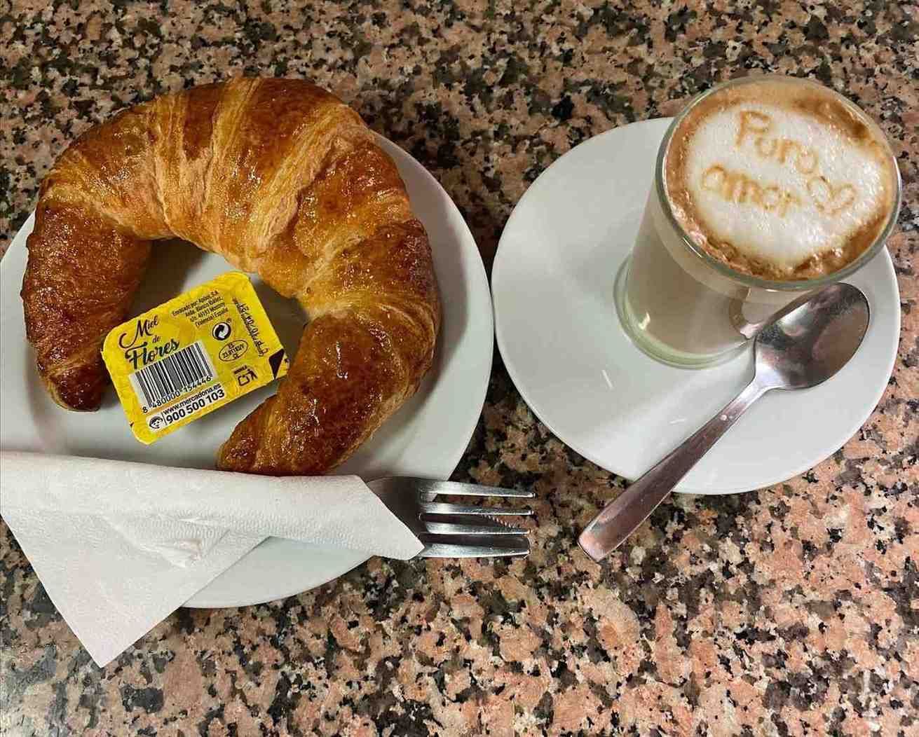 Croissant y capuchino en platillos con cuchara y tenedor sobre una mesa de café moteada.
