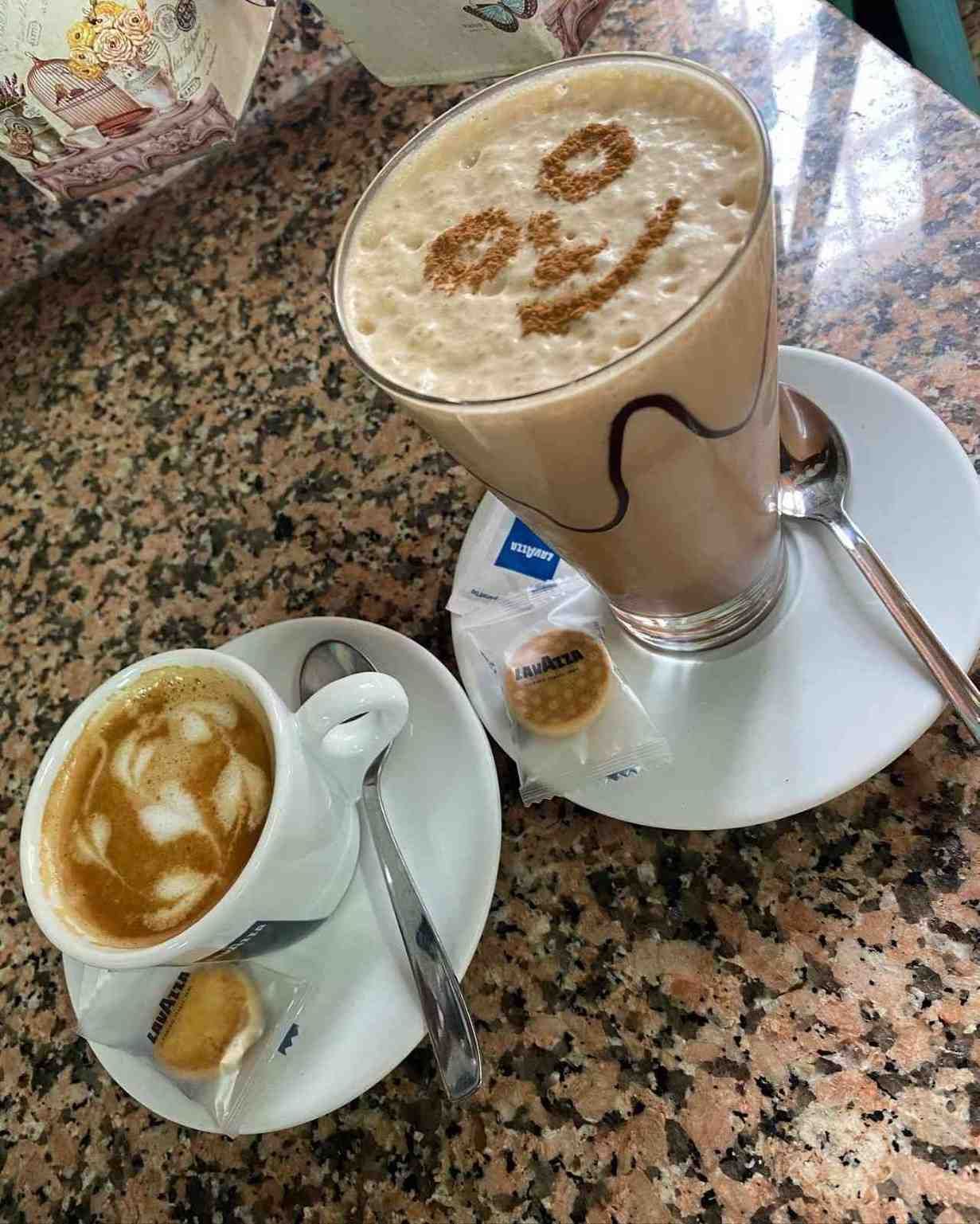 Dos tazas de café en sus platillos, una en un vaso alto con una decoración de espuma sobre una mesa de granito.