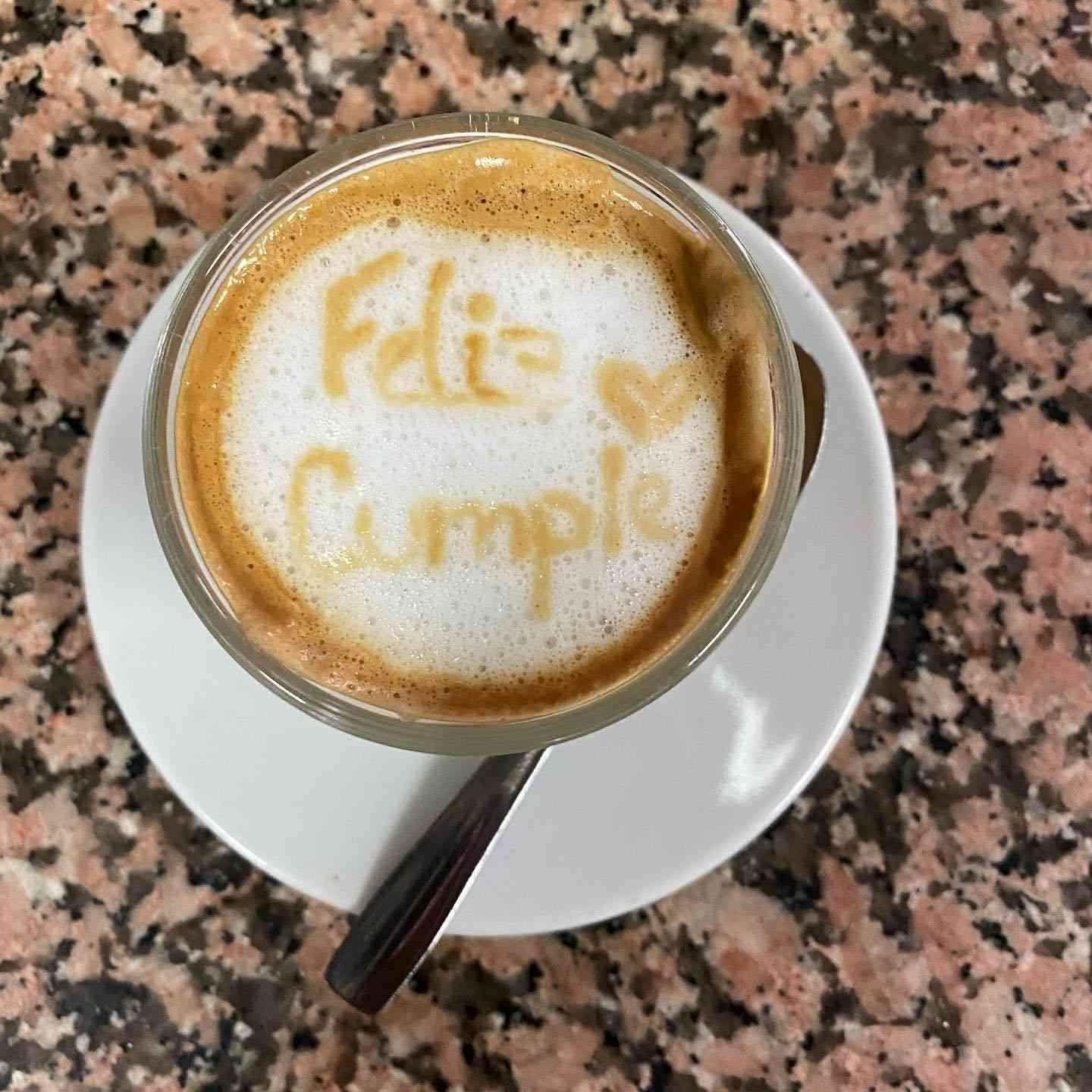 Taza de café sobre platillo con dibujo de latte art que dice “Feliz Cumple”, sobre una mesa de granito