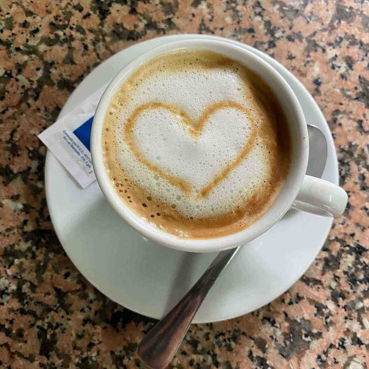 Capuchino con arte latte en forma de corazón en una taza blanca sobre un platillo con una cuchara.