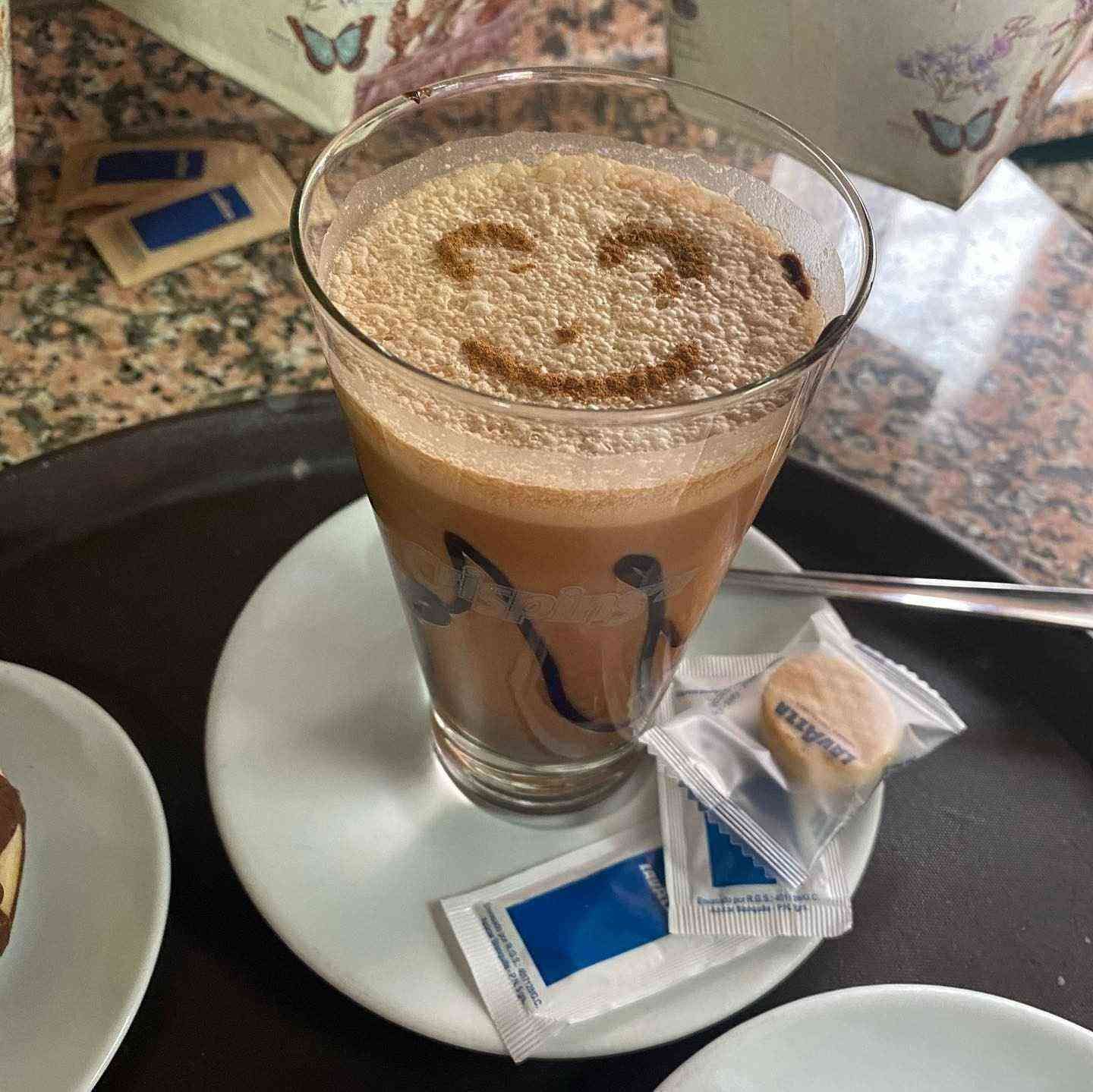 Vaso de café espumoso con un dibujo de una cara hecha con canela en un platillo, junto a sobres de azúcar en una mesa.