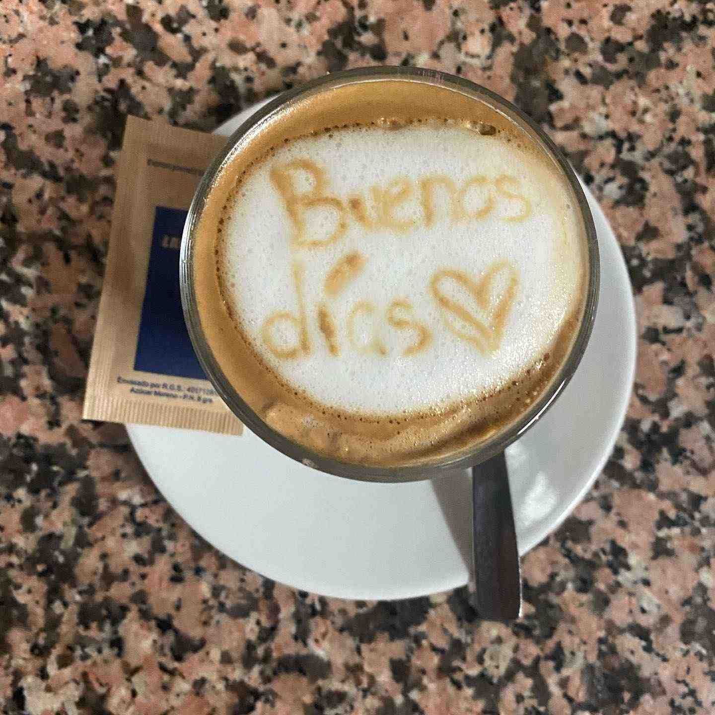 Taza de café con un dibujo en espuma que dice “Buenos días” en un platillo con sobres de azúcar sobre una mesa de granito