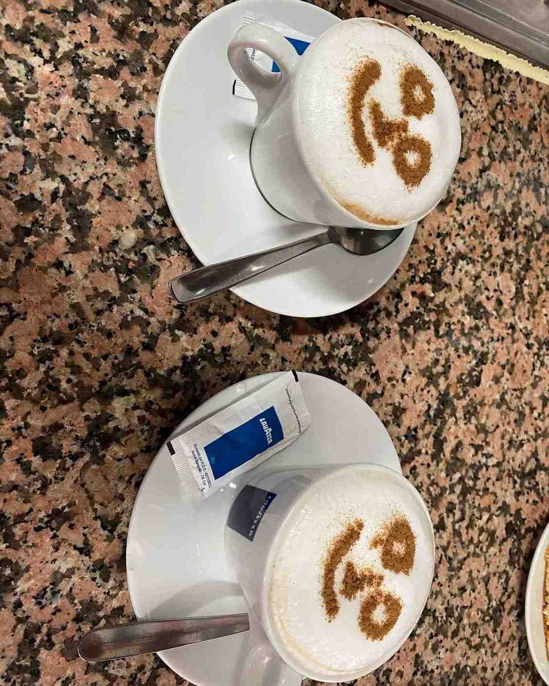 Dos capuchinos blancos en sus platillos sobre una mesa de café moteada, cada uno con su cuchara y sobre de azúcar.