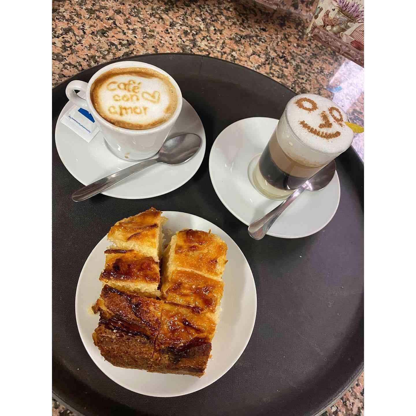 Café y bollería sobre una mesa: capuchino, café con leche y un plato de rebanadas de postre hojaldrado.