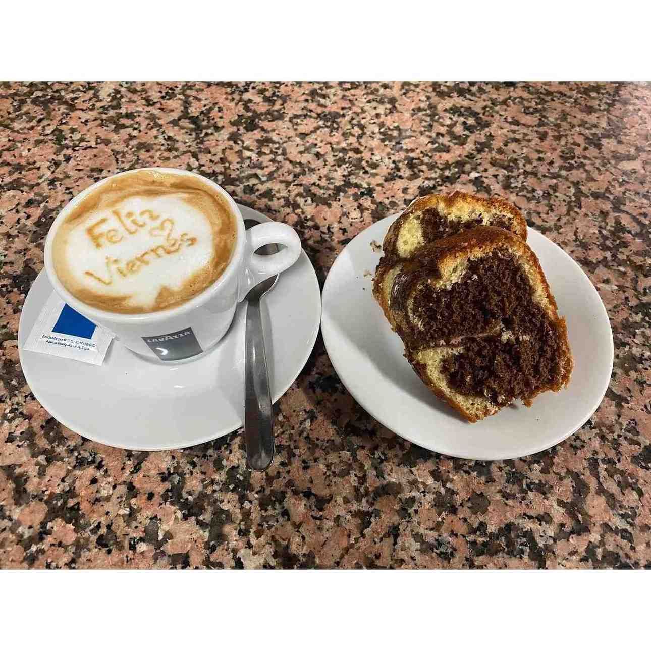 Taza de café con arte latte junto a una rebanada de pastel de mármol en un platillo, sobre una encimera moteada.