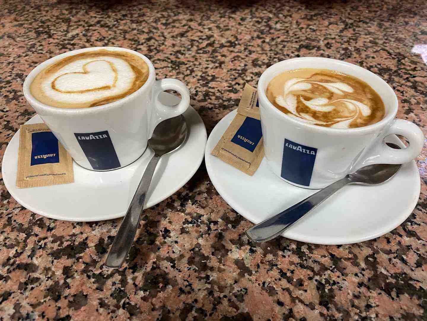 Dos tazas de café con leche en sus platillos con cucharas sobre una mesa de granito moteado.