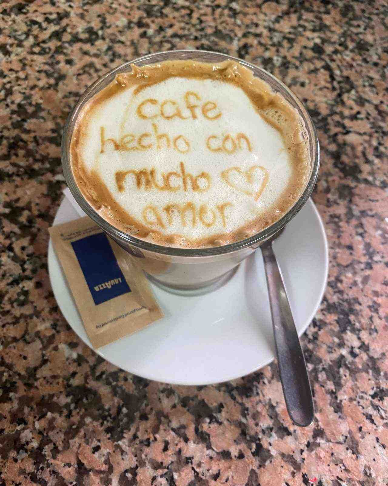 Latte con espuma que dice “café hecho con mucho amor”, en platillo con sobre de azúcar y cuchara.