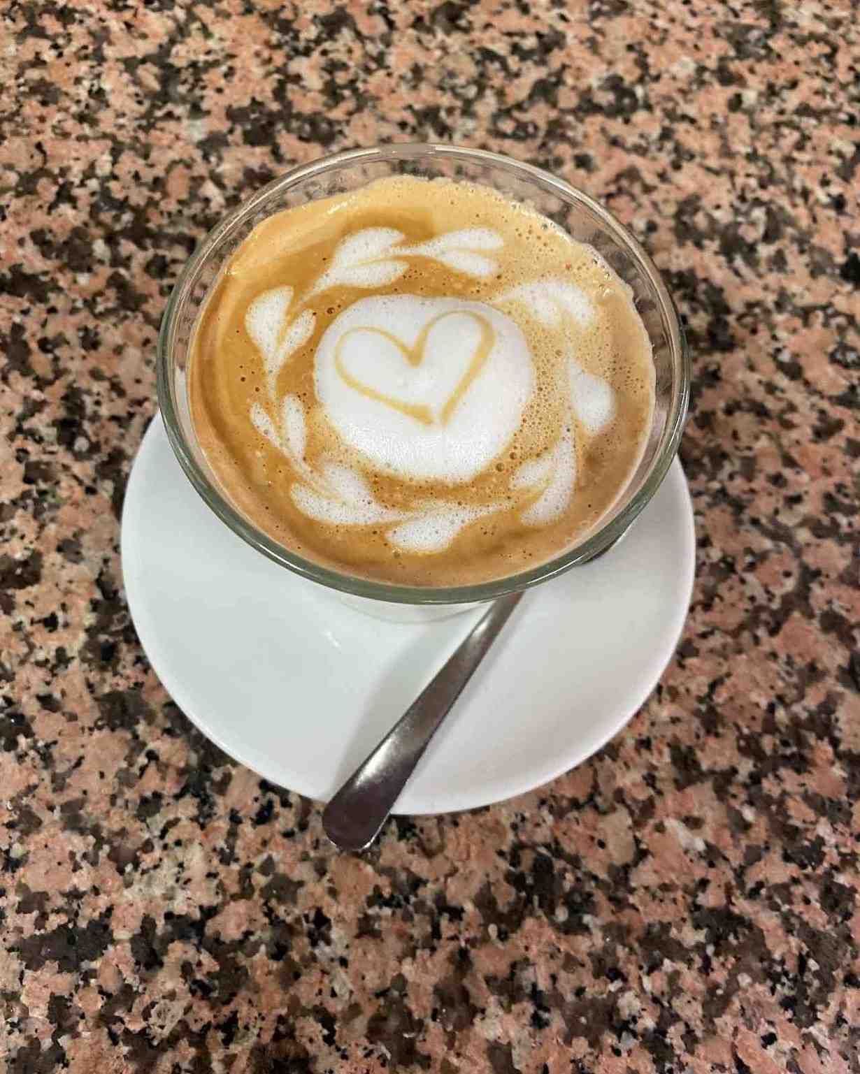 Café con leche decorado con espuma en forma de corazón sobre un platillo blanco con una cuchara, sobre una mesa moteada.
