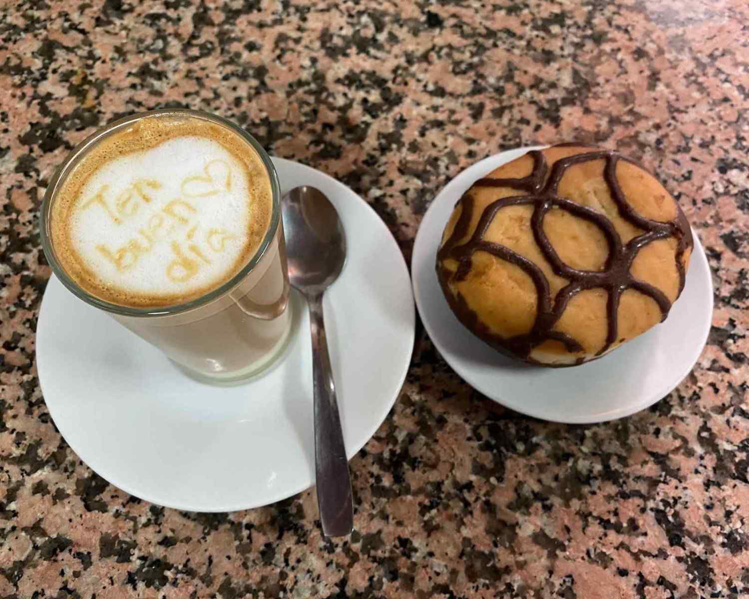 Taza de café con leche con espuma en forma de corazón junto a un pastelito bañado en chocolate sobre un plato blanco.