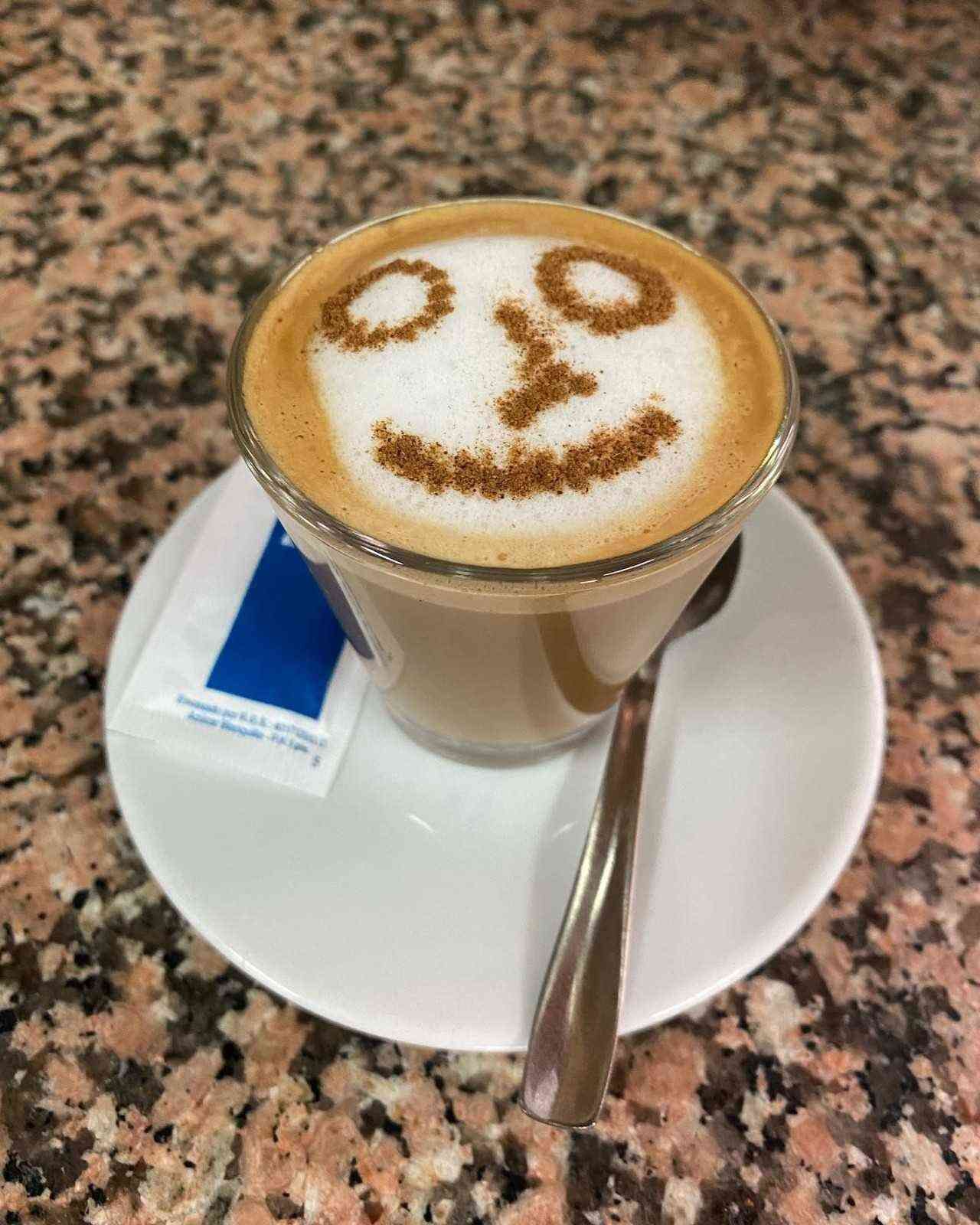 Capuchino con espuma decorada con una carita sonriente en un platillo con cuchara y sobre de azúcar.