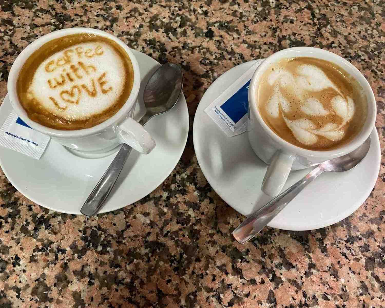 Dos capuchinos en platillos con cucharas, uno con dibujos de latte art y el otro con la inscripción 