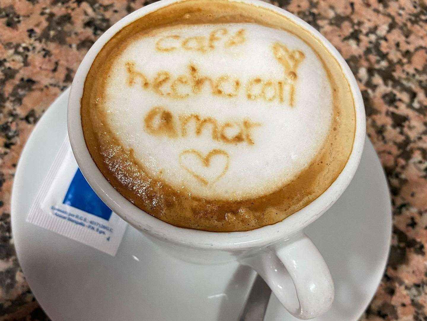 Latte en una taza y platillo blancos con un dibujo en la espuma que dice “hecho con amor” y un corazón.