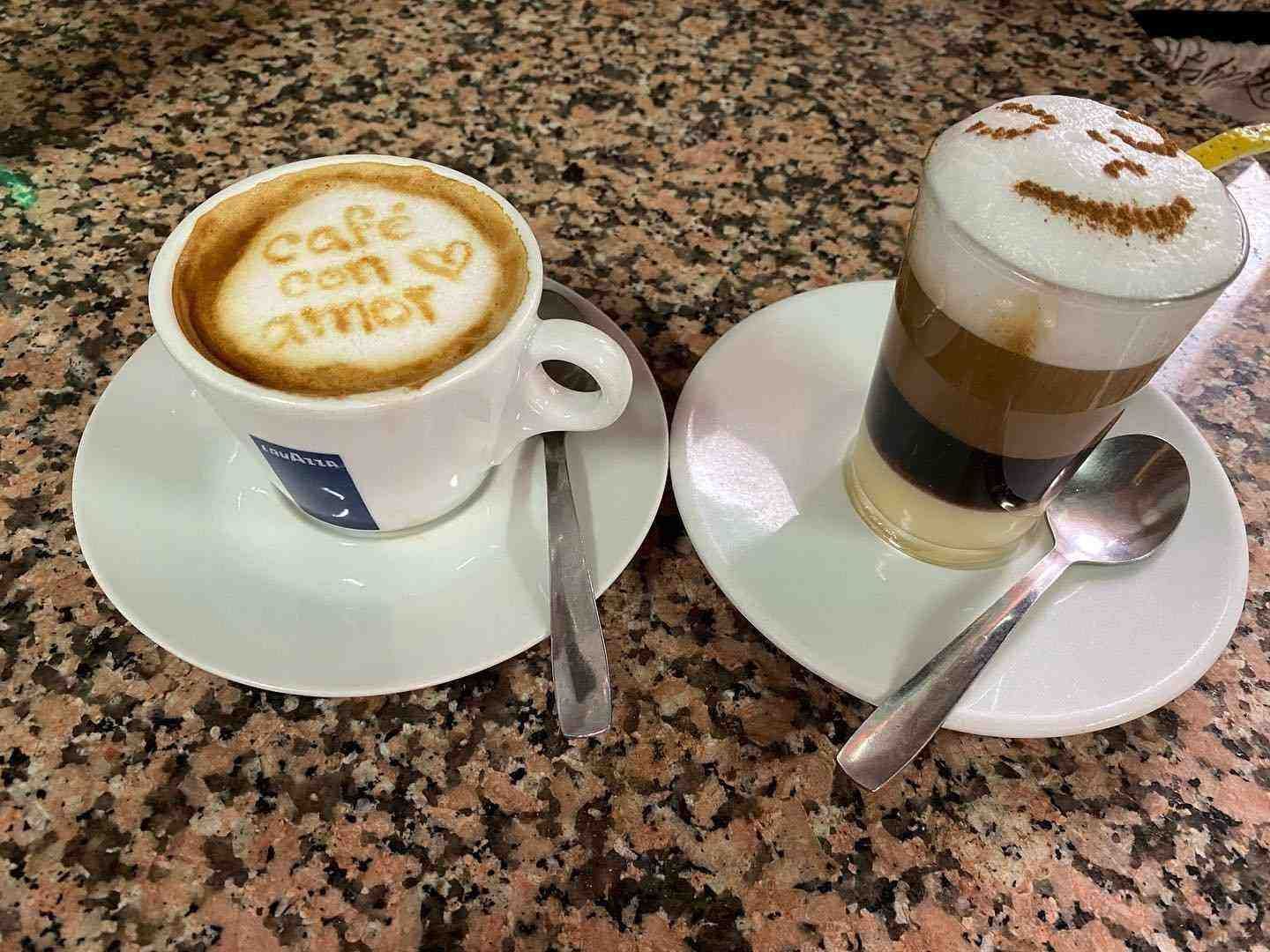 Dos capuchinos en platillos sobre una mesa moteada, cada uno con figuras de espuma y cucharas a su lado.