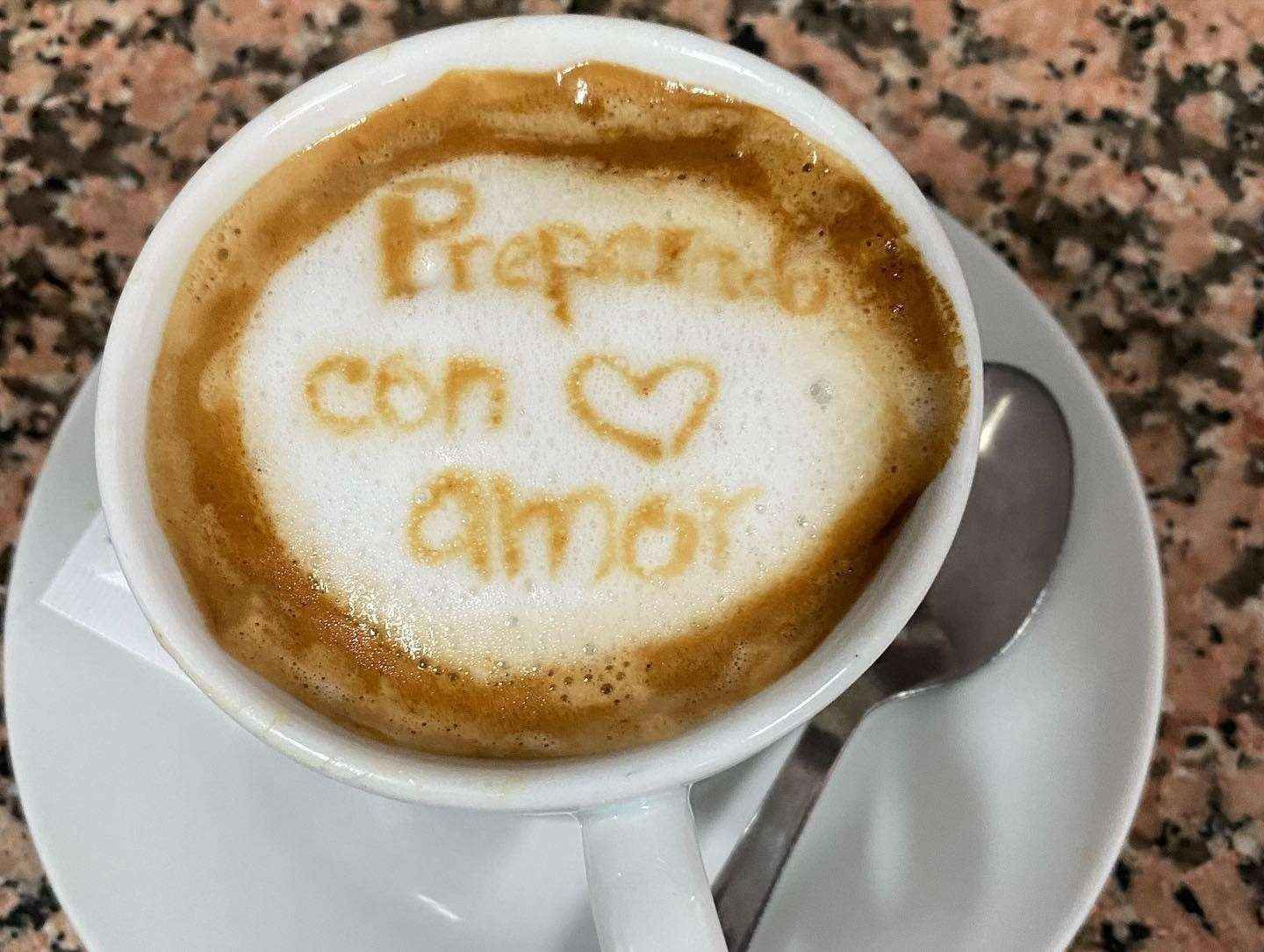 Latte con un dibujo en la espuma que dice “Prepárate con amor” en un platillo blanco con una cuchara.