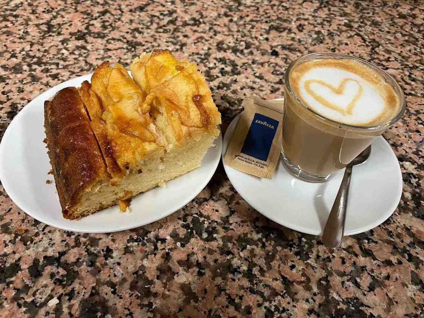 Un trozo de pastel y un café con leche decorado con arte latte en pequeños platos blancos sobre una mesa moteada.