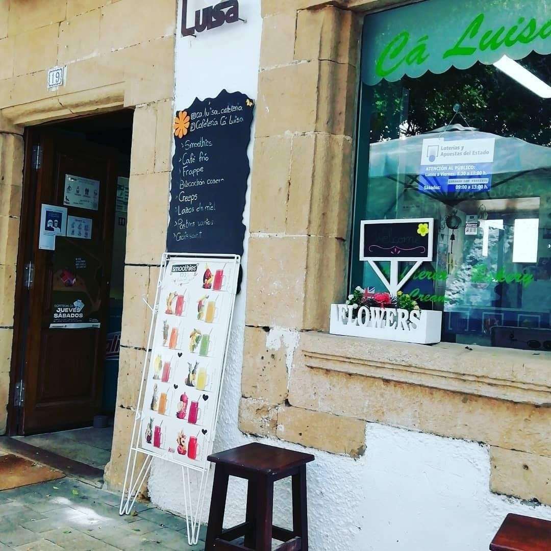 Pequeña cafetería con la puerta abierta, pizarra con el menú, escaparate y un taburete de madera en el exterior.
