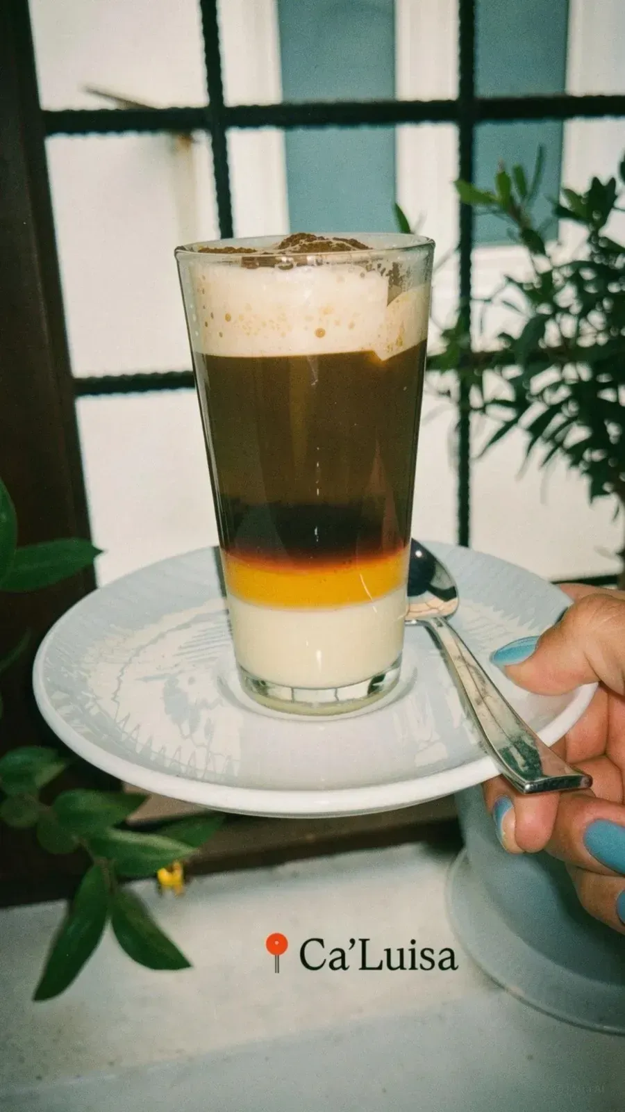 Bebida de café en capas servida en un vaso transparente sobre un platillo, con una mano sosteniendo una cuchara al lado.