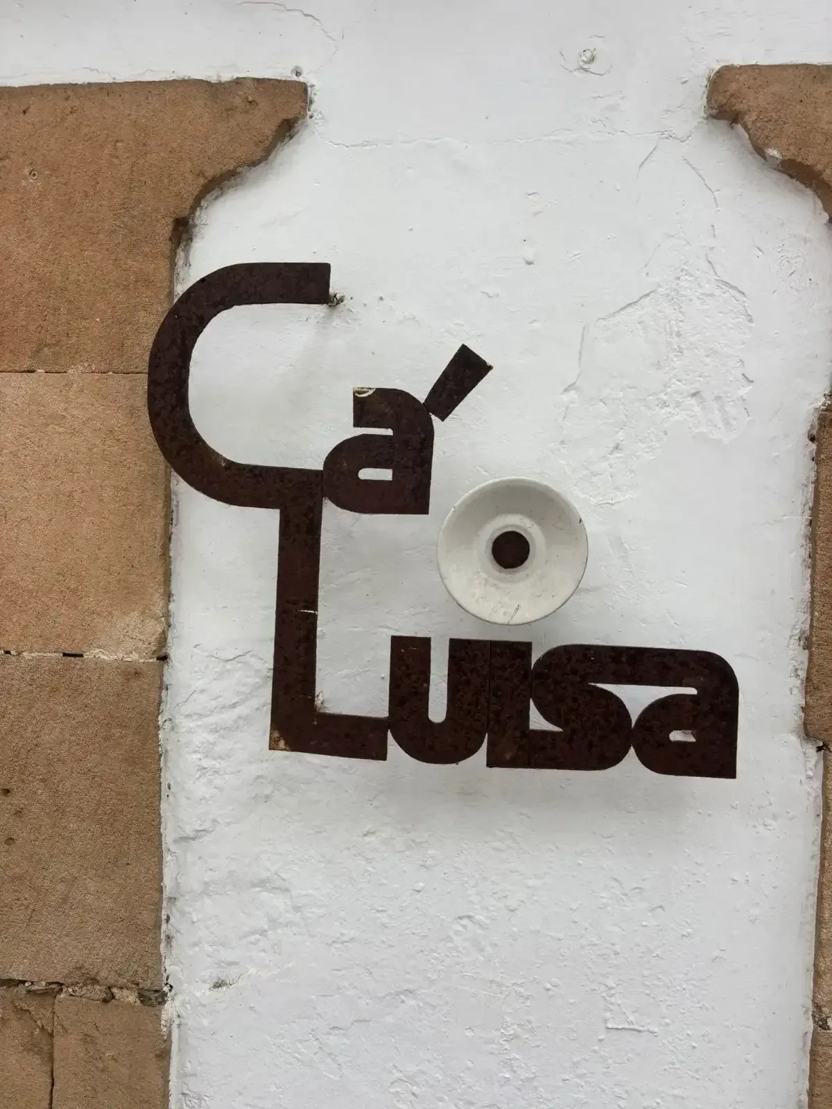 Letrero marrón “Ca de Luisa” en una pared blanca con una lámpara redonda.