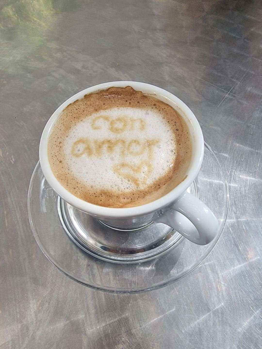 Taza de café con leche decorada con espuma sobre un platillo encima de una mesa de acero inoxidable.