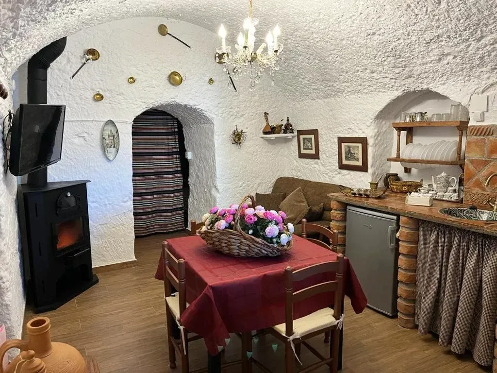 Interior de una casa cueva con comedor, chimenea y cocina. Paredes encaladas, piedra vista y detalles en rojo.