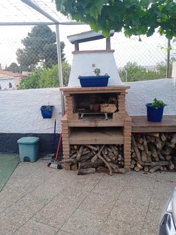 Parrilla de ladrillo con leñero, pared blanca y plantas.
