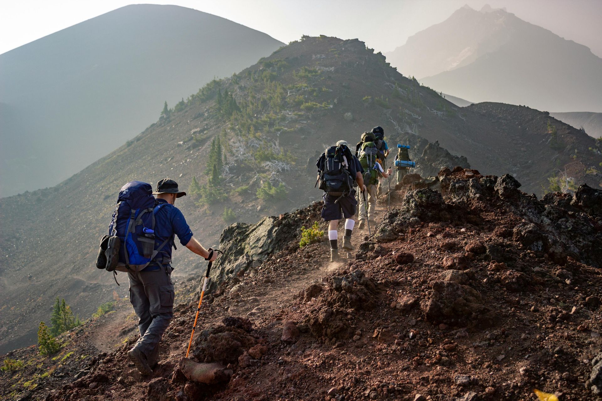 Excursionistas con mochilas ascienden por un sendero rocoso de montaña bajo un cielo brumoso.