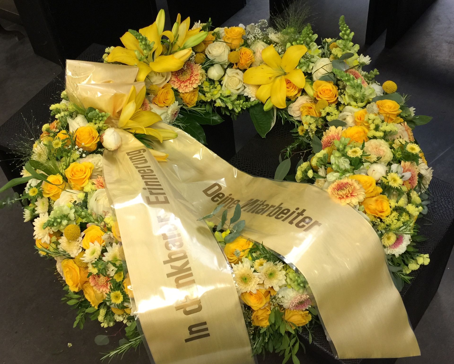 Ein Kranz aus gelben und weißen Blumen mit einem goldenen Band, auf dem steht: in dankbarer Erinnerung