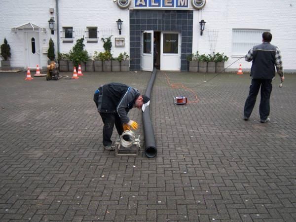 Zwei Männer arbeiten an einem Rohr vor einem Gebäude mit der Aufschrift