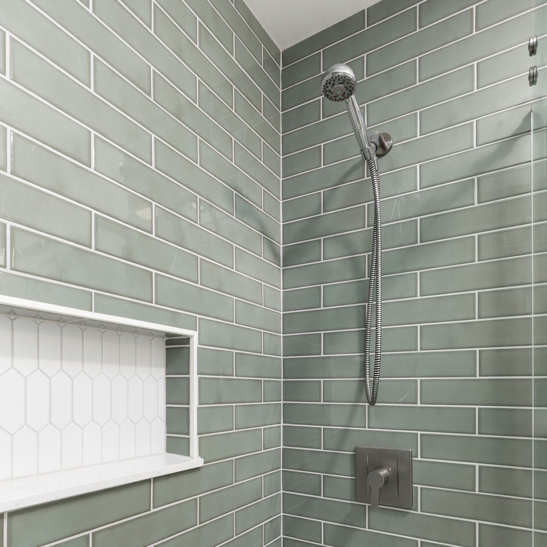 Douche avec murs en carrelage métro vert, étagère encastrée blanche et pomme de douche argentée.