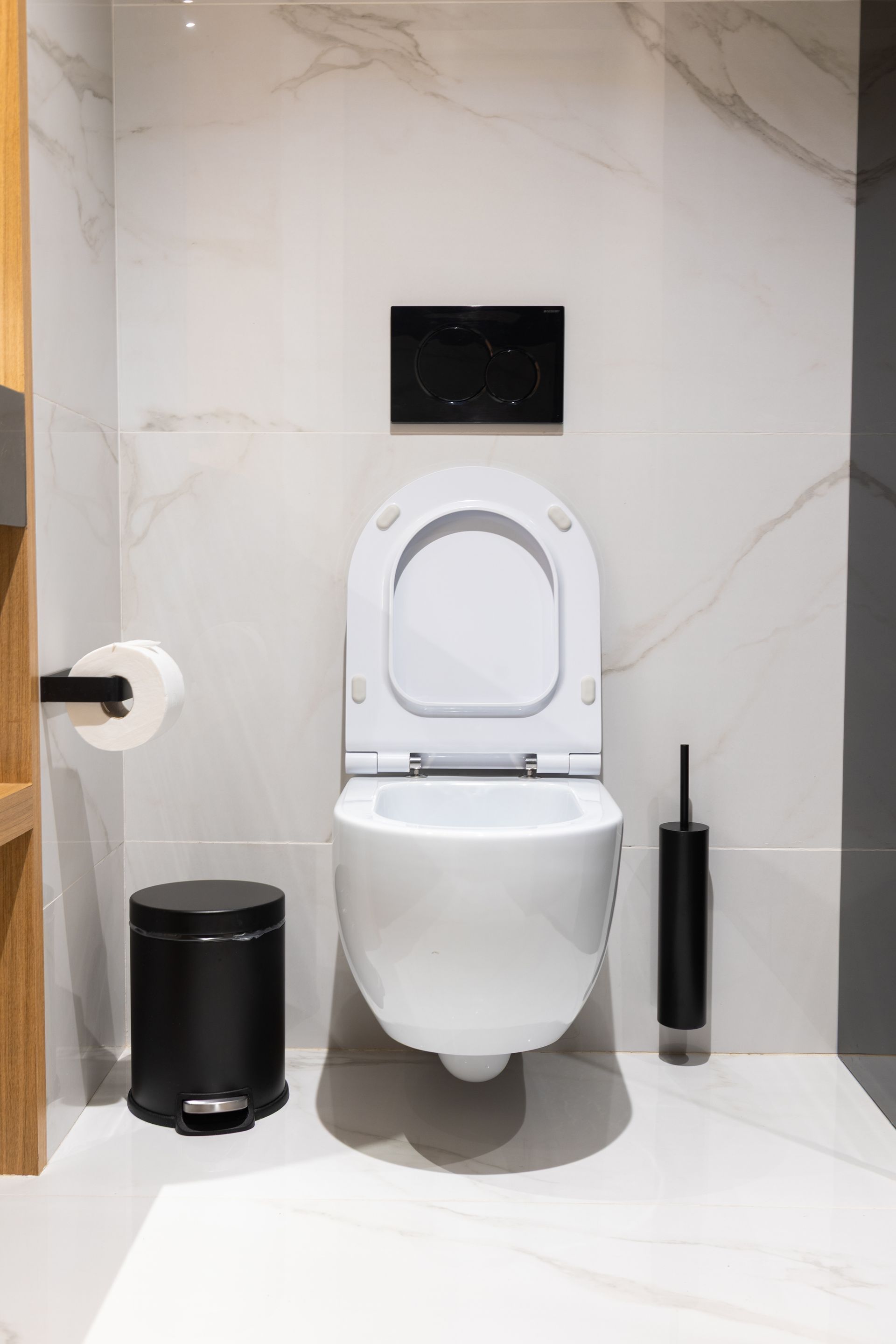 Toilettes murales avec abattant ouvert, plaque de chasse d’eau et porte-papier toilette, contre un mur en marbre.