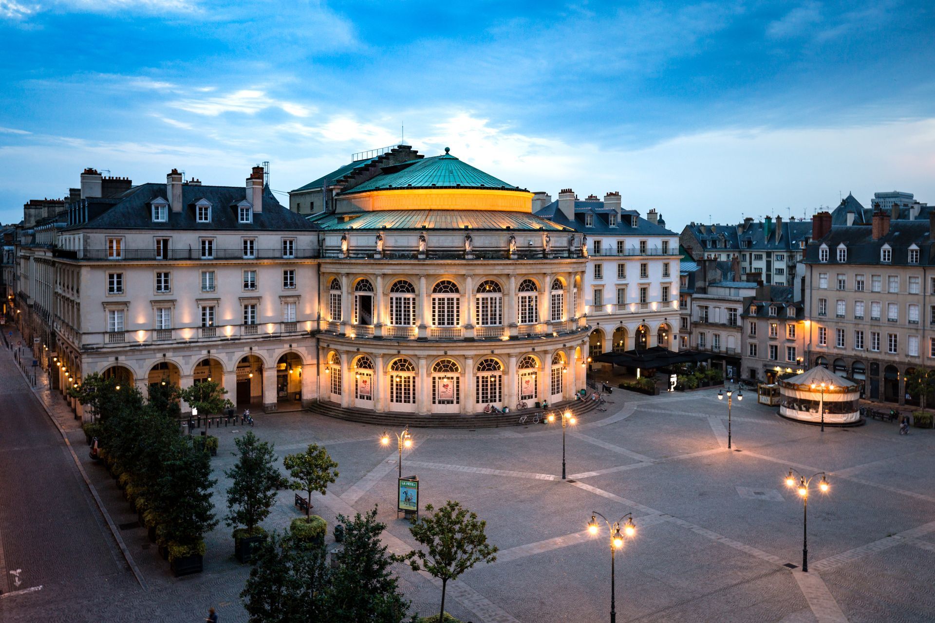 Un grand théâtre éclairé avec un dôme en cuivre sur une place de la ville au crépuscule.