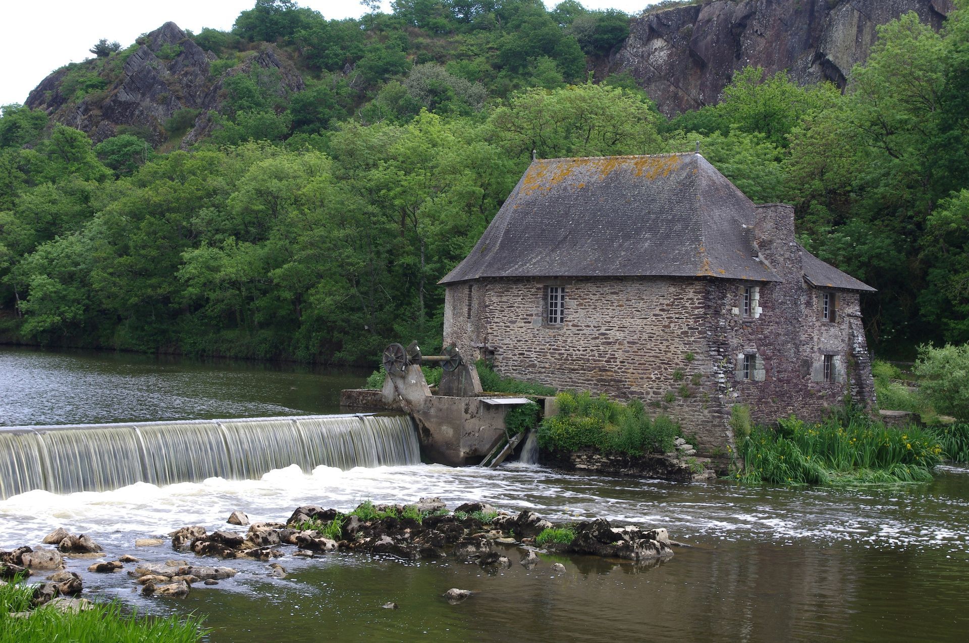 Moulin en pierre sur une rivière avec une cascade, entouré d'arbres et d'une colline rocheuse.